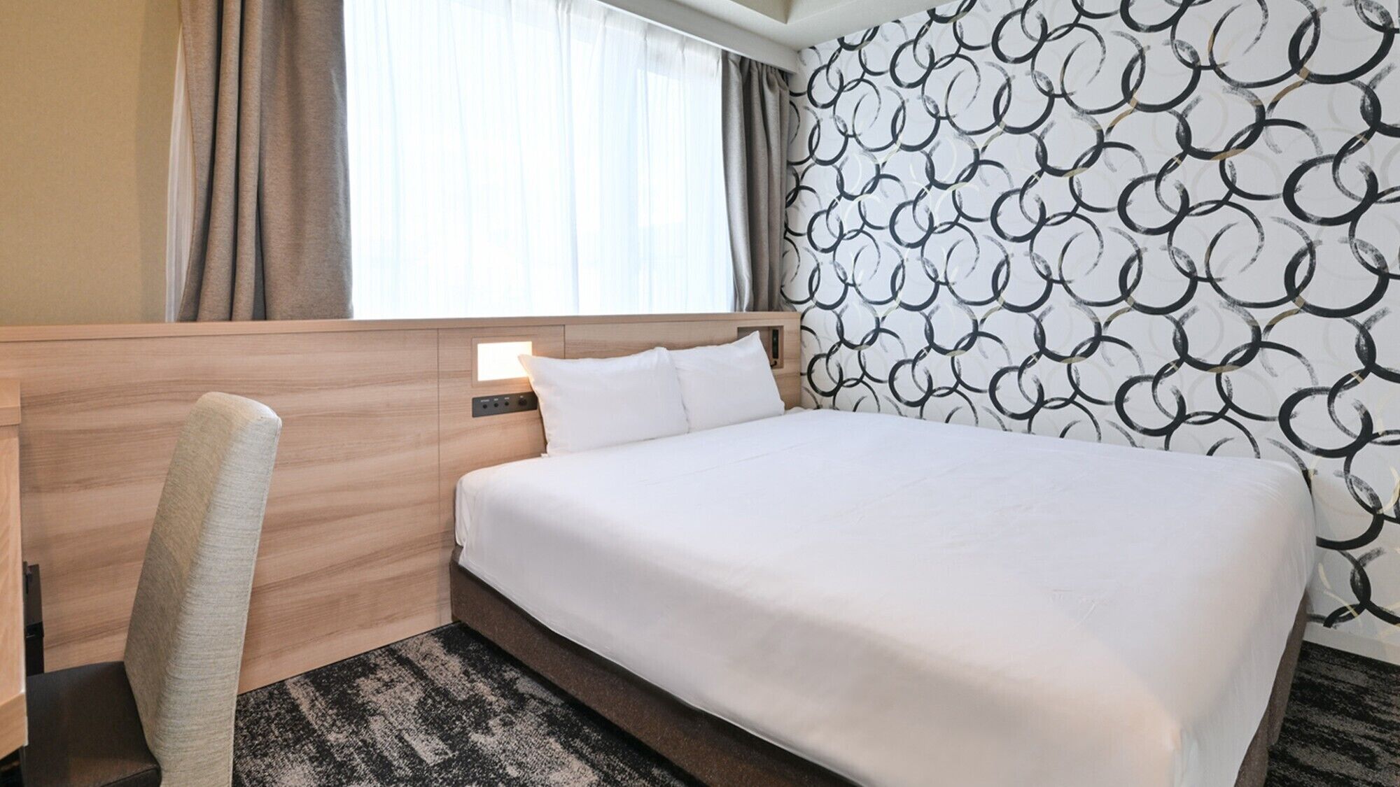 A hotel room with a bed, a chair, a headboard, a window with curtains, and a patterned wall.