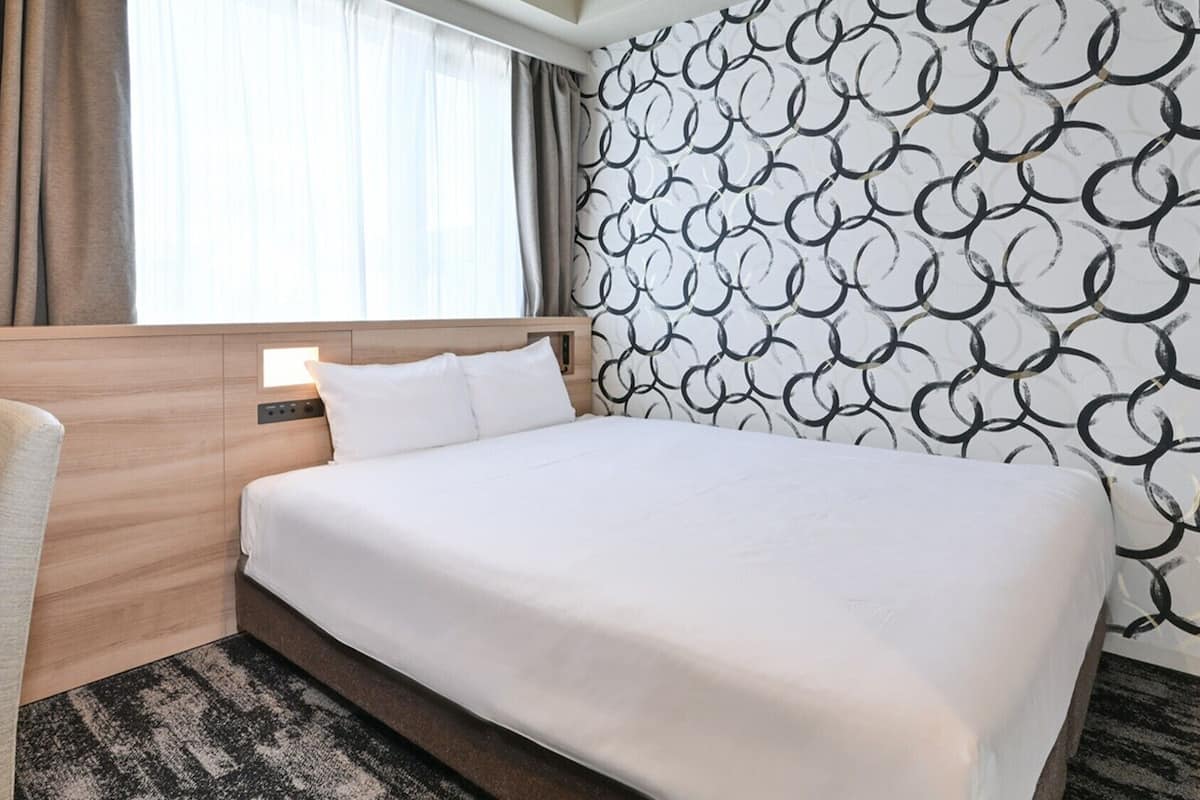 A hotel room with a bed, a chair, a headboard, a window with curtains, and a patterned wall.