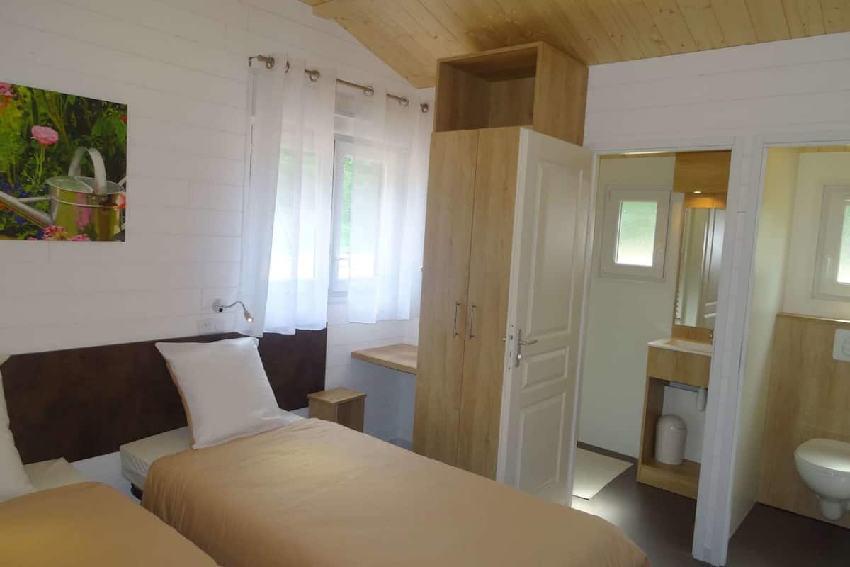 A bedroom with two beds, a wooden wardrobe, a bathroom with a toilet and sink, and a painting of flowers on the wall.