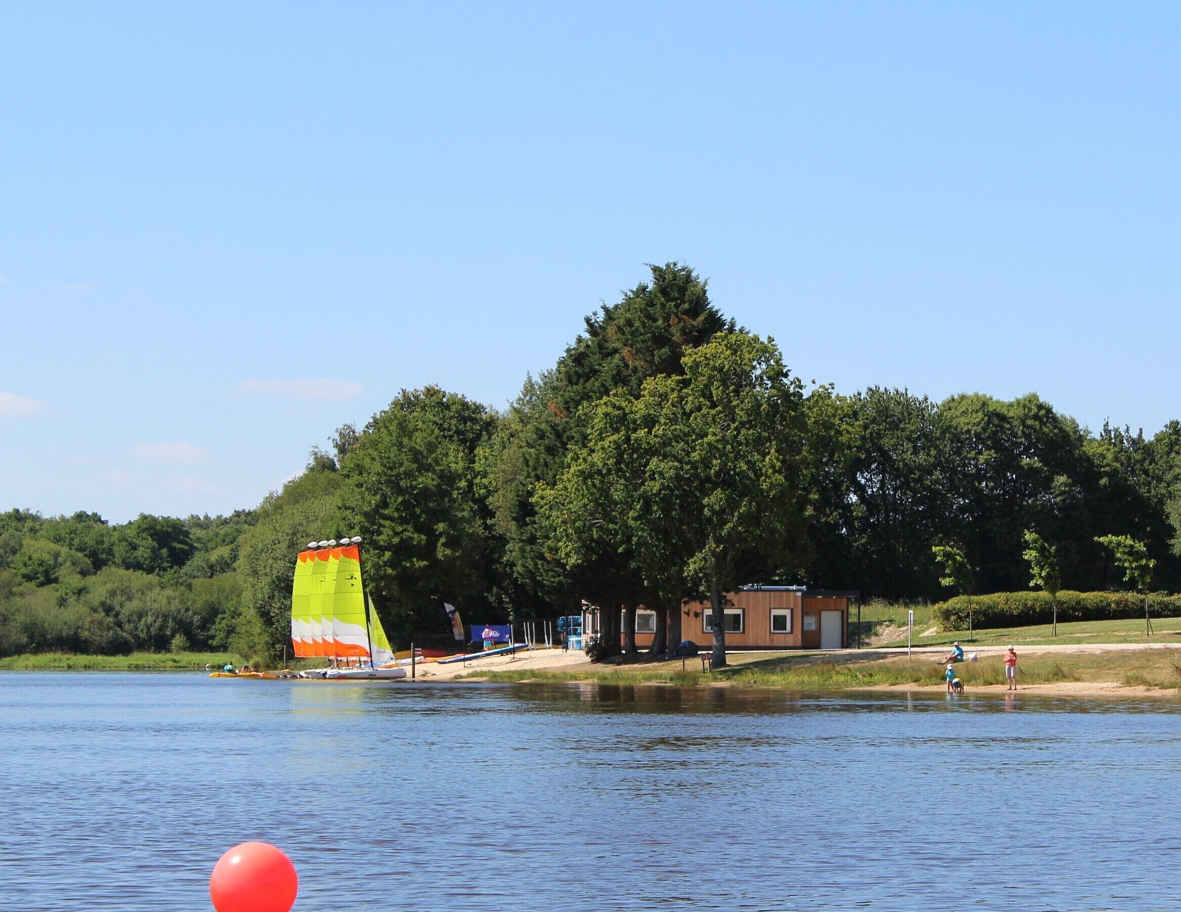 Vista de un lago con un velero, un pequeño edificio de madera y un área boscosa.
