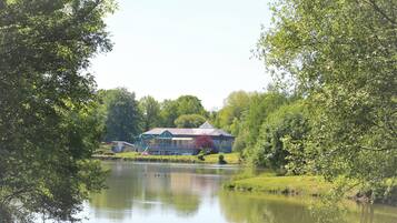 Un bâtiment situé au bord du lac, doté d’une grande terrasse et entouré d’une végétation luxuriante, avec un lac paisible à proximité.