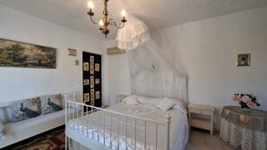 Un dormitorio con una cama blanca, un banco blanco, una mesita de noche, una pequeña mesa con un jarrón de flores y una araña.
