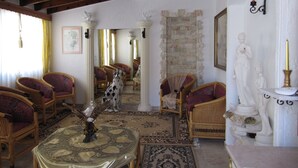 Una habitación con techo alto, pared de piedra y una estatua. Hay sillas de mimbre, una mesa con un pájaro decorativo y un perro.