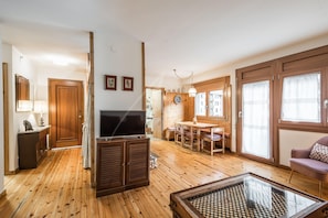 A spacious living area with wooden flooring, a television, a glass coffee table, and a dining table with chairs.