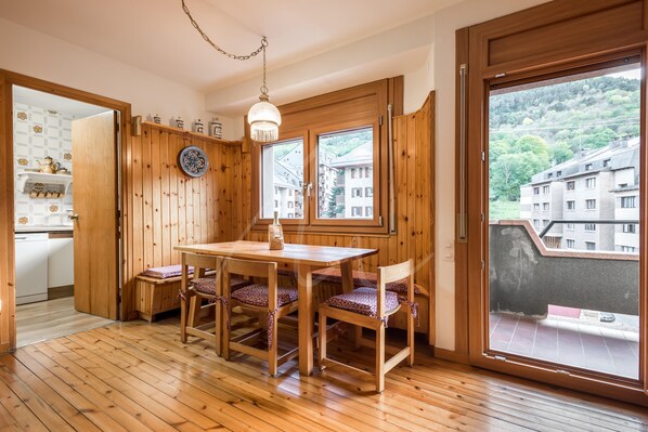 A wooden dining area with a table and chairs, large windows with a view of buildings and greenery, and a kitchen visible through an open door.