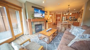A cozy living room with a fireplace, a large TV, and a kitchen in the background.