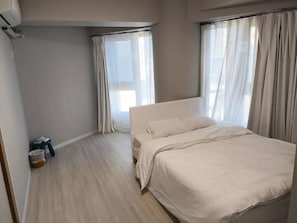 A bedroom with a bed, two windows with curtains, a small table with items, and a trash bin.