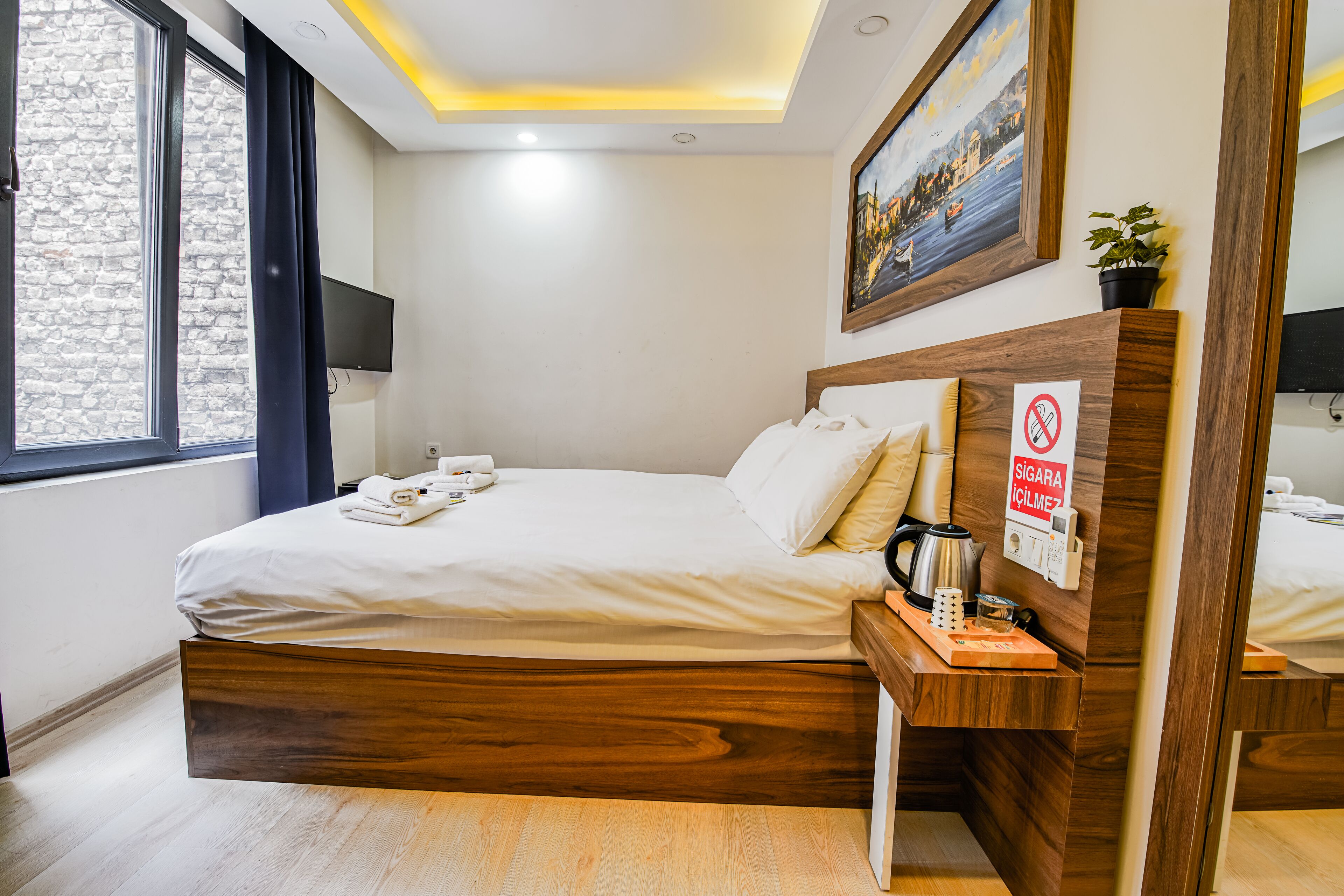 Ein modernes Hotelzimmer mit einem hölzernen Bett, einem Nachttisch mit Kaffeemaschine, einem Bild an der Wand und einem Fenster mit Vorhängen.
