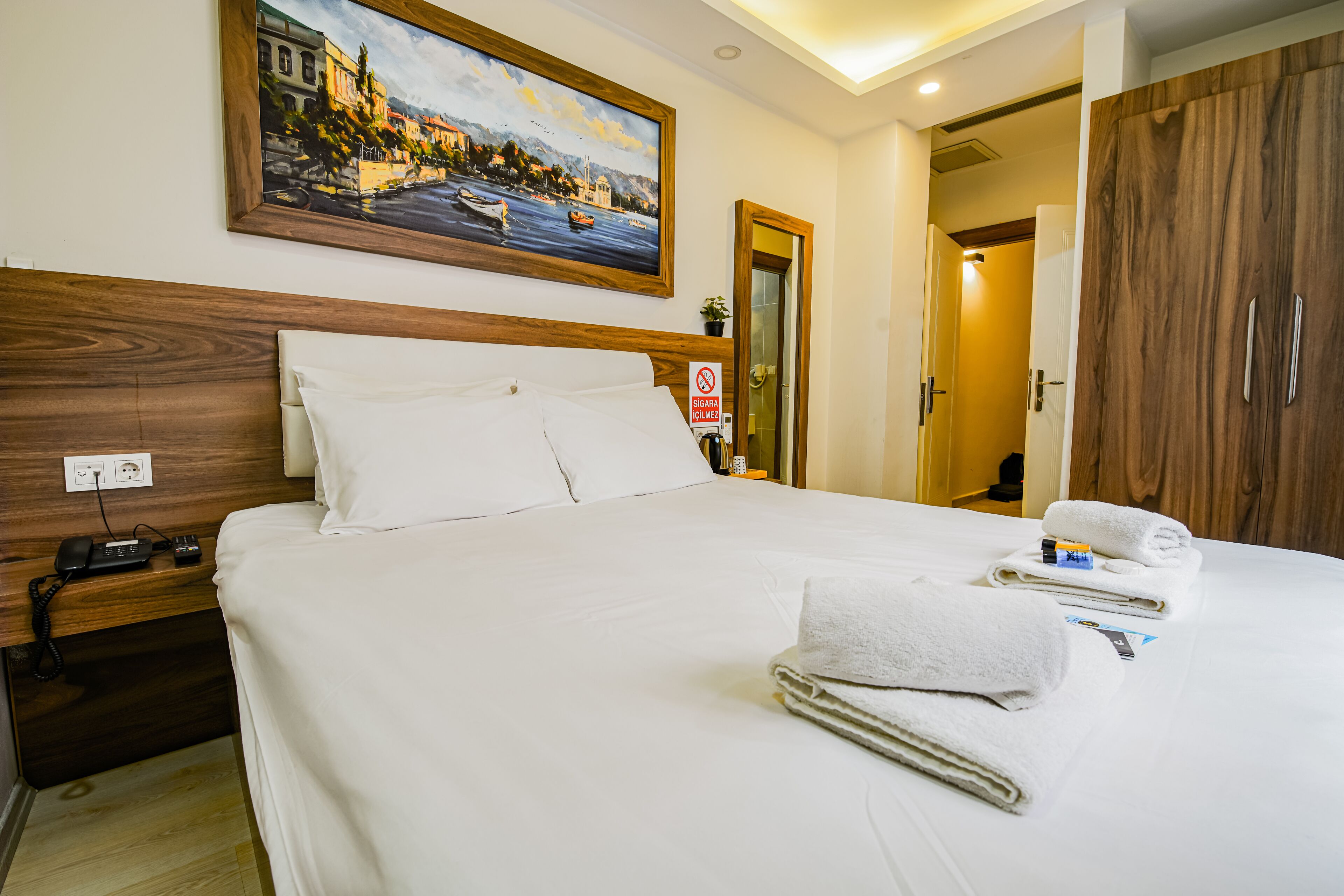 A neatly made bed with white linens, a painting of a coastal scene above it, and a wooden wardrobe to the right.