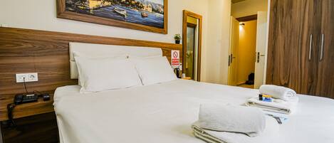 A neatly made bed with white linens, a painting of a coastal scene above it, and a wooden wardrobe to the right.