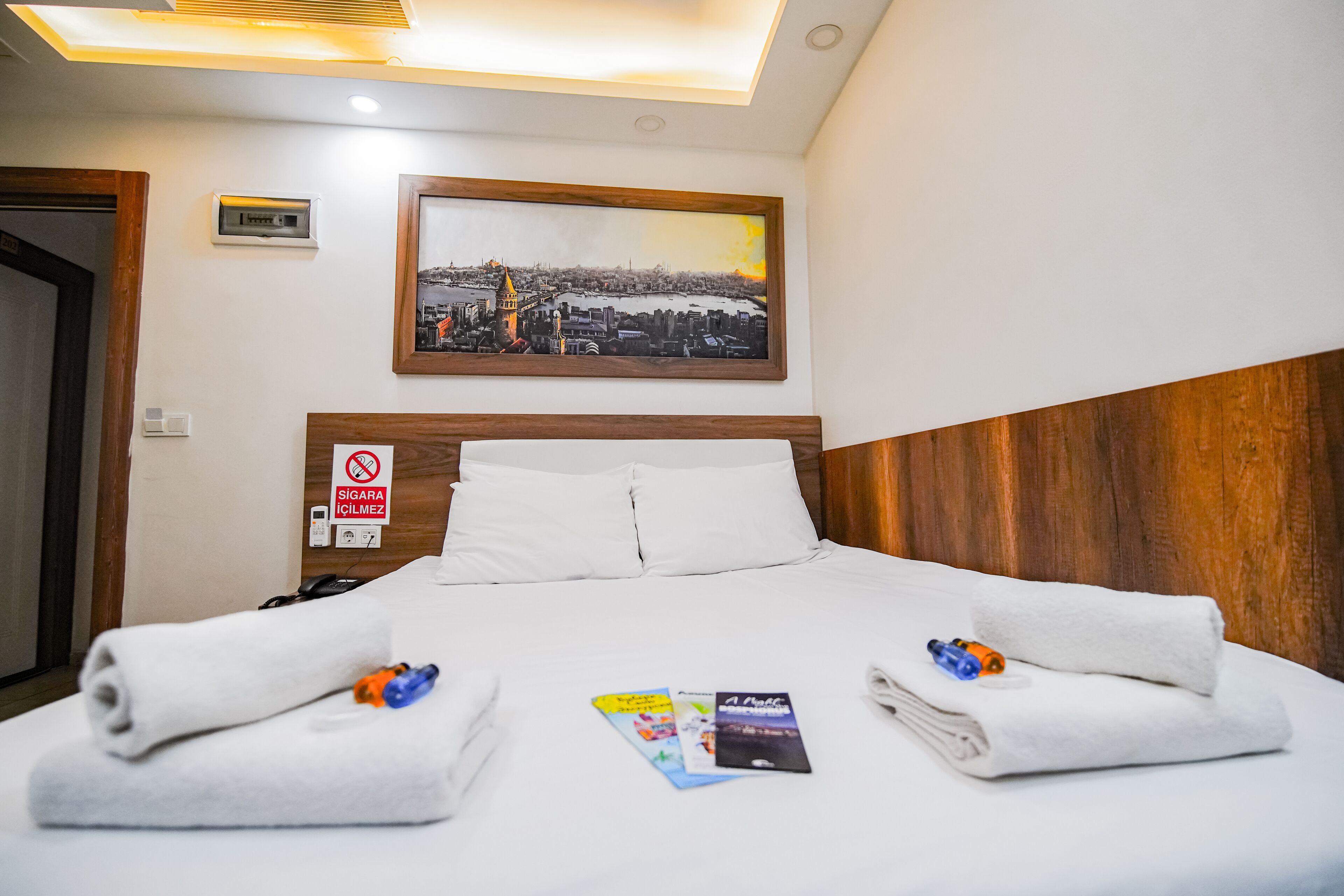 Une chambre d’hôtel avec un lit parfaitement fait, un tableau représentant une vue de la ville encadré au-dessus de la tête de lit, et un panneau « interdiction de fumer » sur le mur.