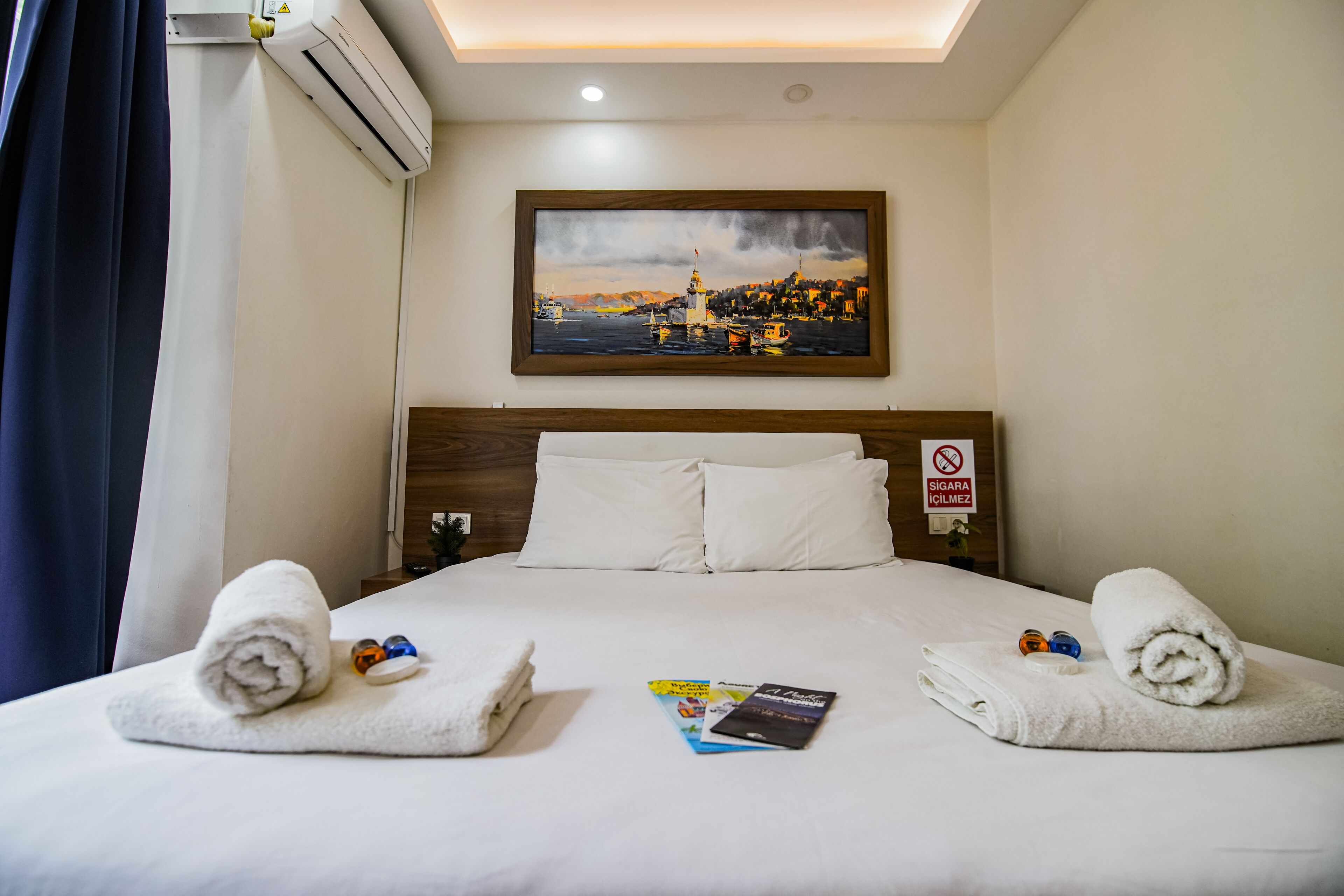 A hotel room with a neatly made bed, a painting above it, and a no-smoking sign on the wall.