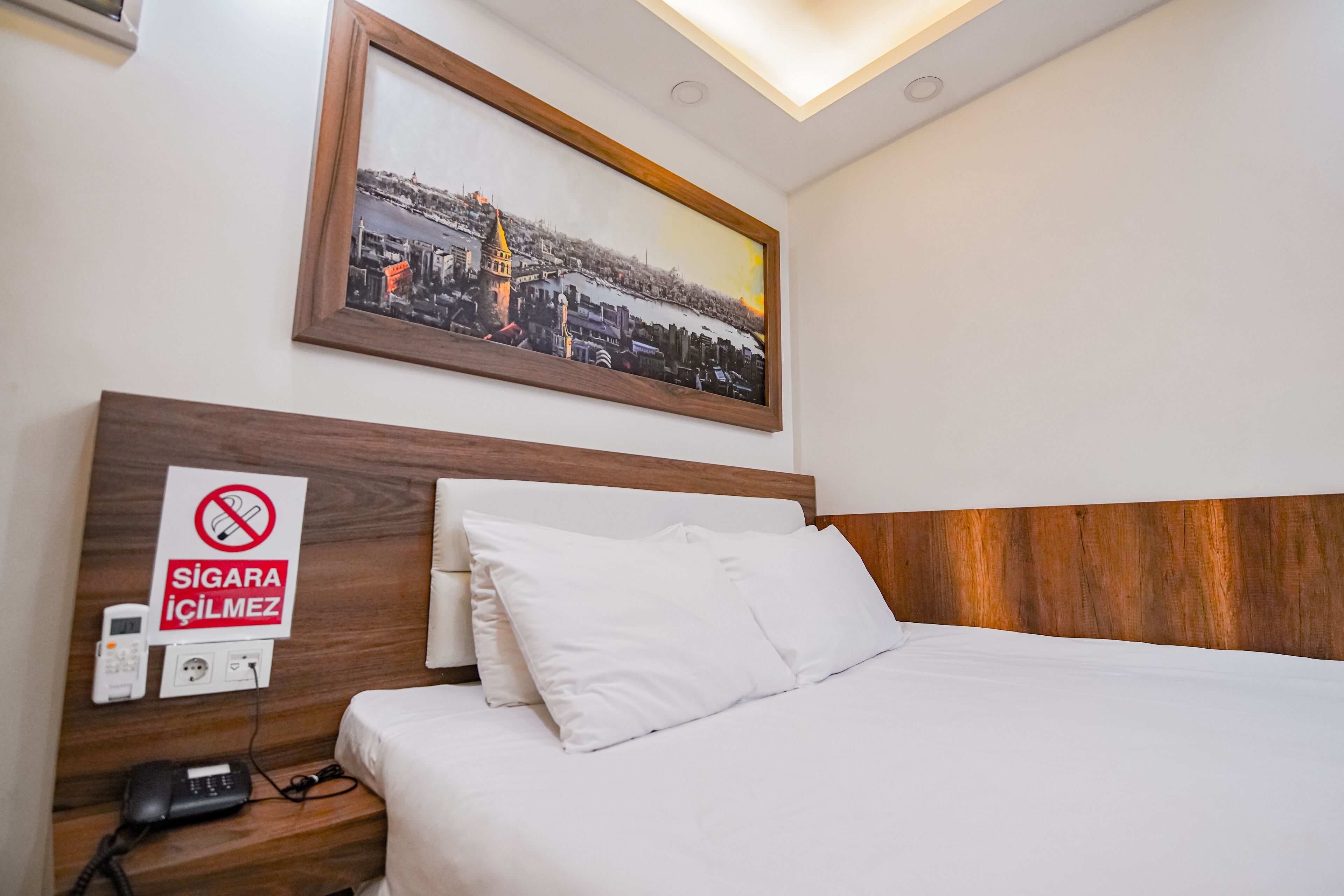 A hotel room with a bed, a nightstand with a phone, a framed picture on the wall, and a sign indicating no smoking.