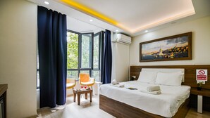 A hotel room with a large bed, a desk, two chairs, a painting, and a view of greenery through the window. - Mitani Suites (Istanbul)