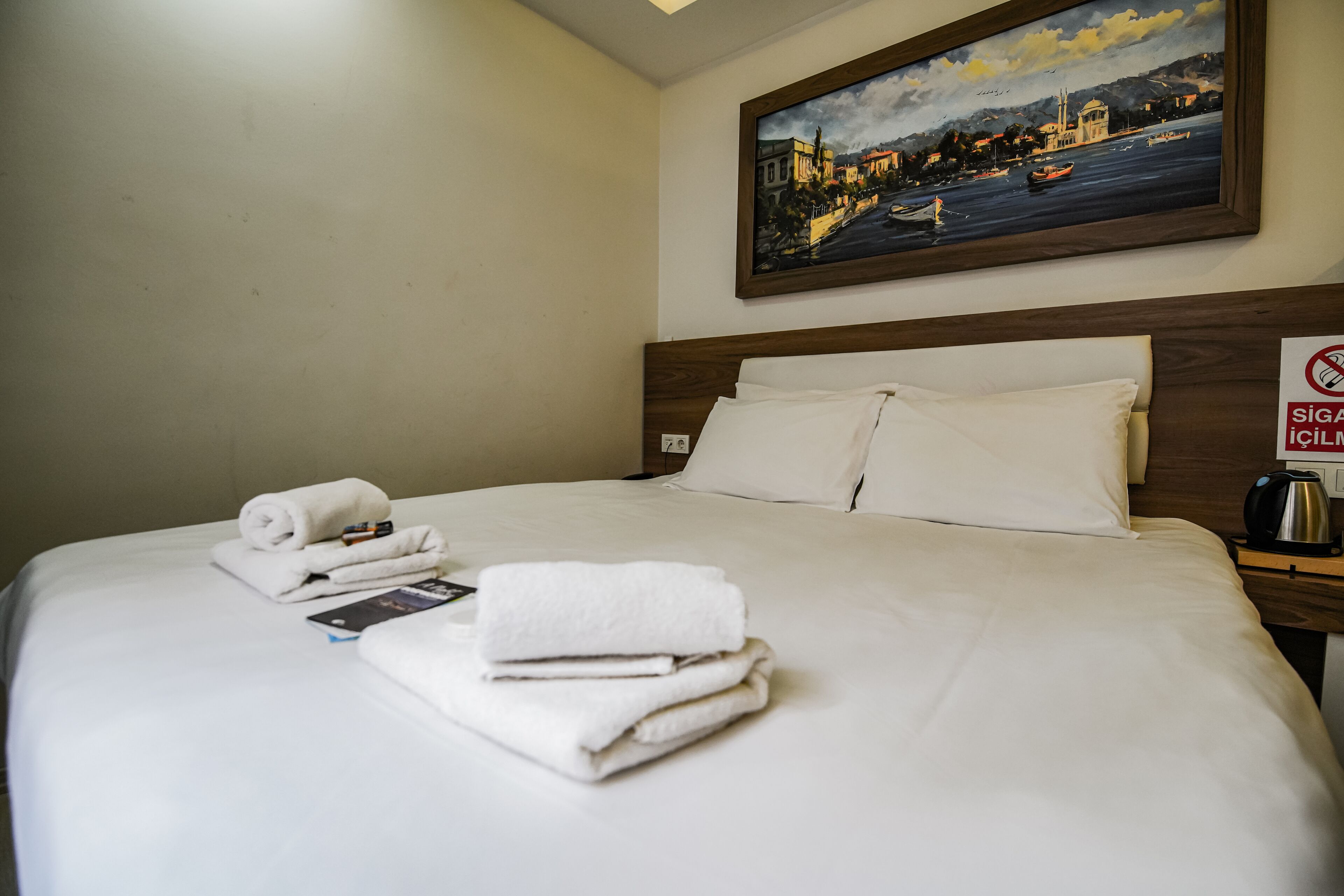 A neatly made bed with folded towels and a book, a painting of a cityscape on the wall, and a sign indicating no smoking.