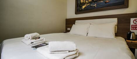 A neatly made bed with folded towels and a book, a painting of a cityscape on the wall, and a sign indicating no smoking.