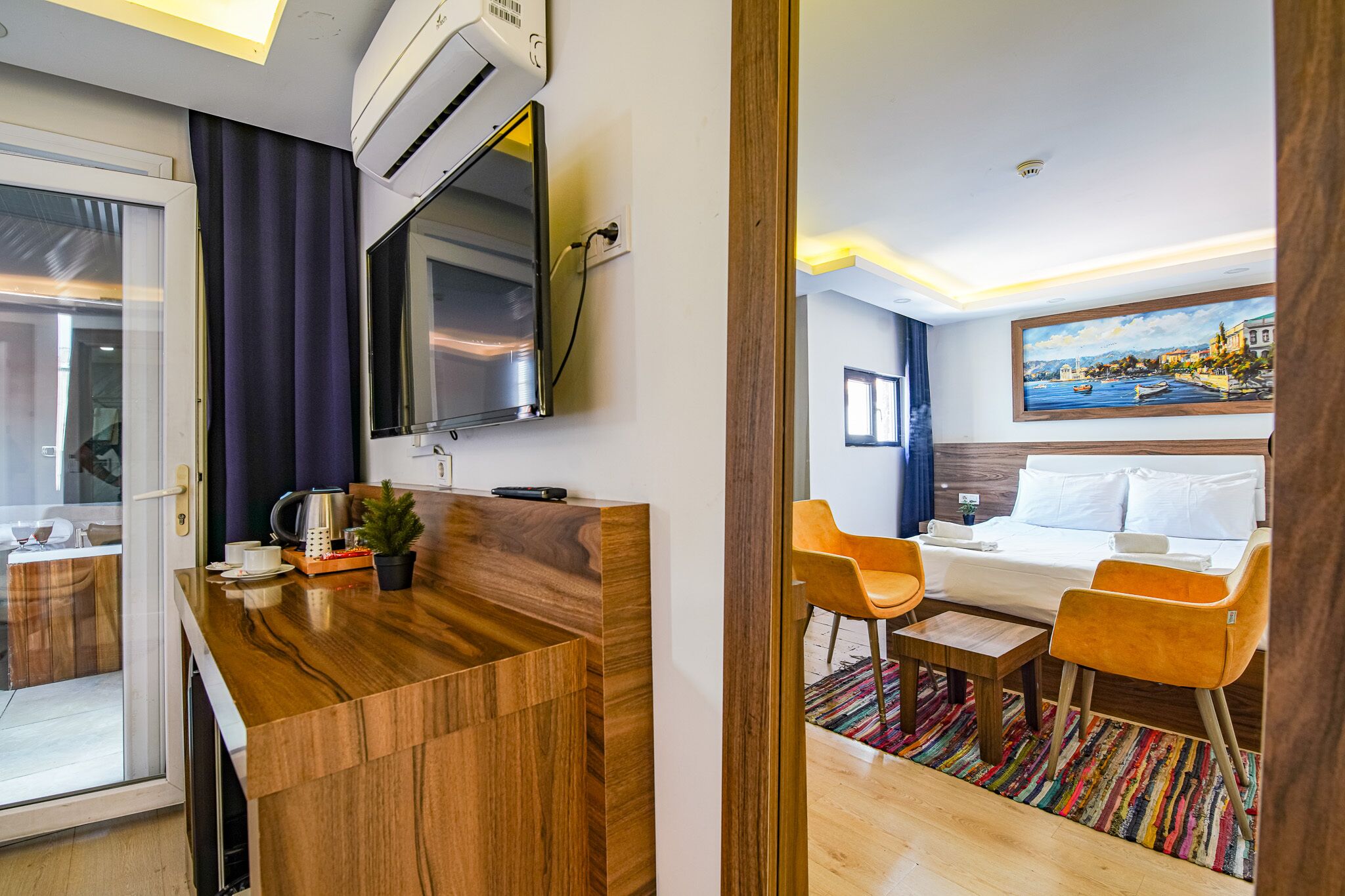 A hotel room with a wooden desk, a bed with two orange chairs, a painting of a coastal town, and a view of the sea through the window.