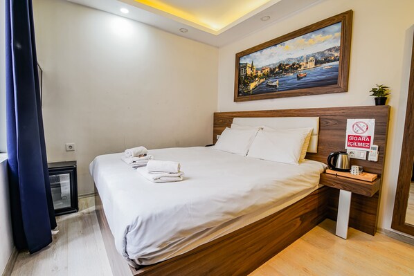 A neatly made bed with white linens and a folded towel, a wooden headboard, a nightstand with a coffee maker, and a framed painting of a coastal scene.