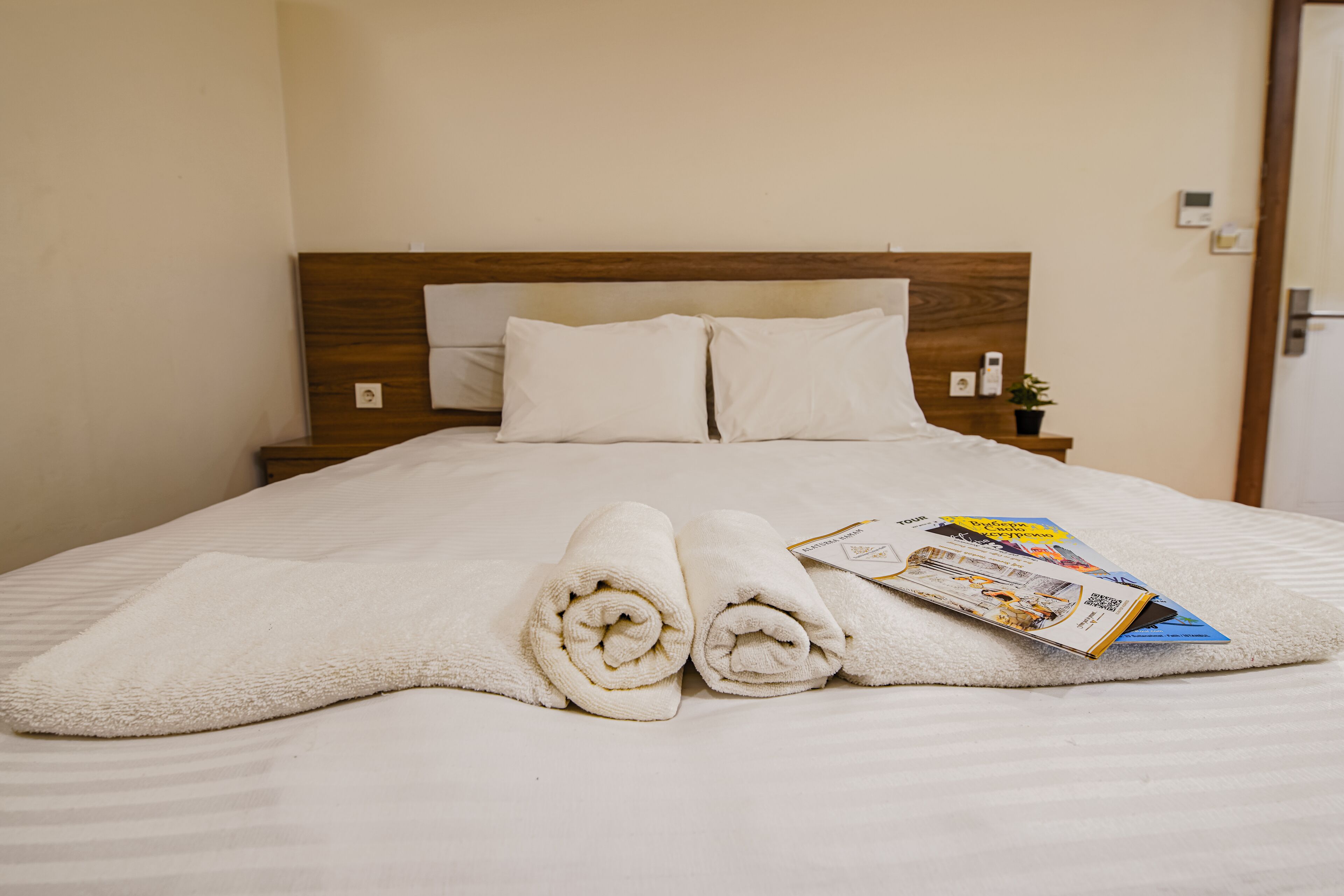 A neatly made bed with white linens and rolled towels, a wooden headboard, and a small bedside table with a plant.