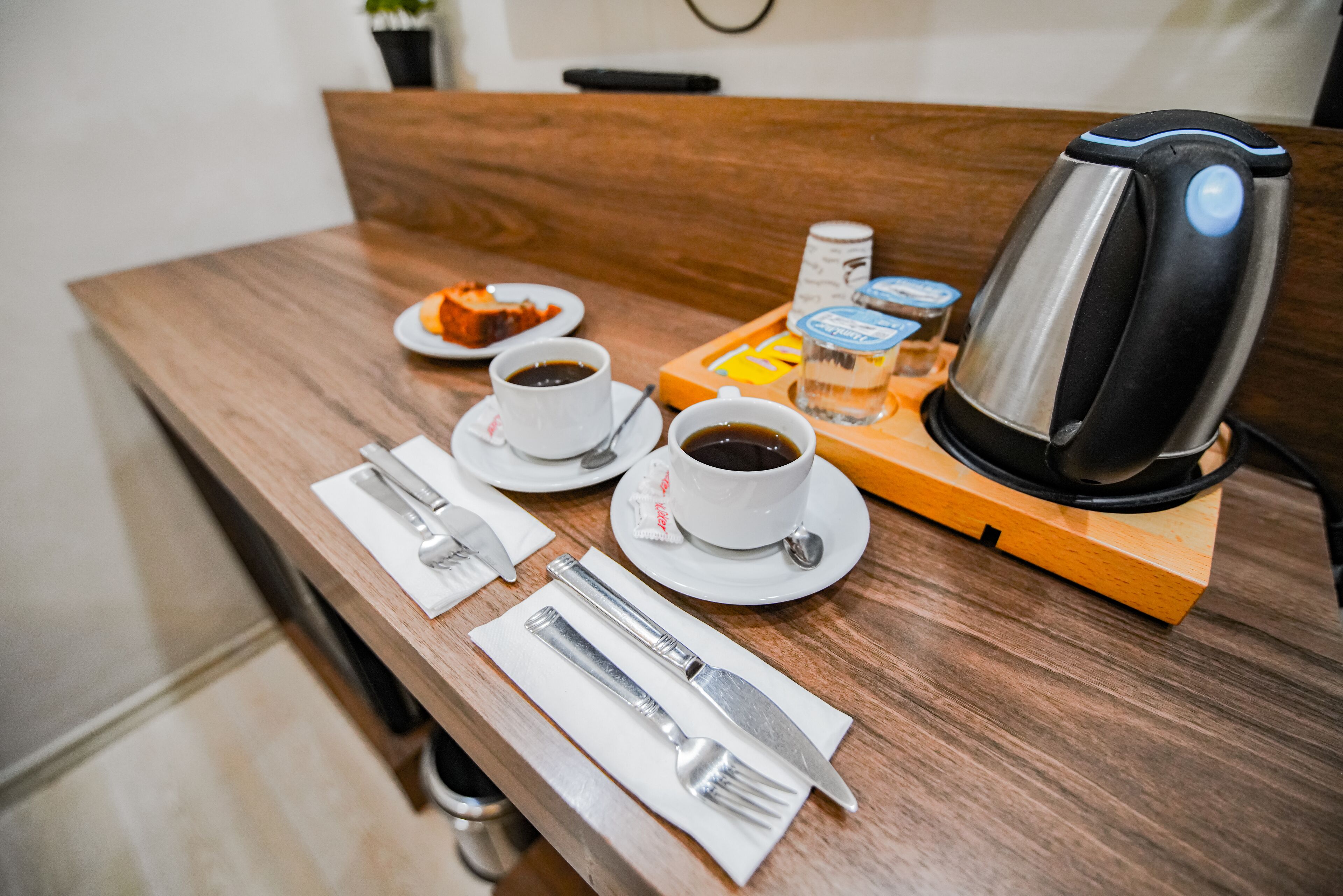 A wooden table with a coffee maker, two cups of coffee, a plate of food, and a tray with condiments.