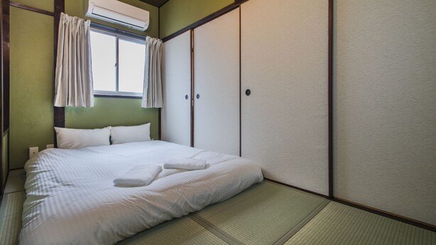 Un lit bien fait, avec du linge de lit blanc, une fenêtre avec des rideaux, un appareil de climatisation et une porte coulissante.