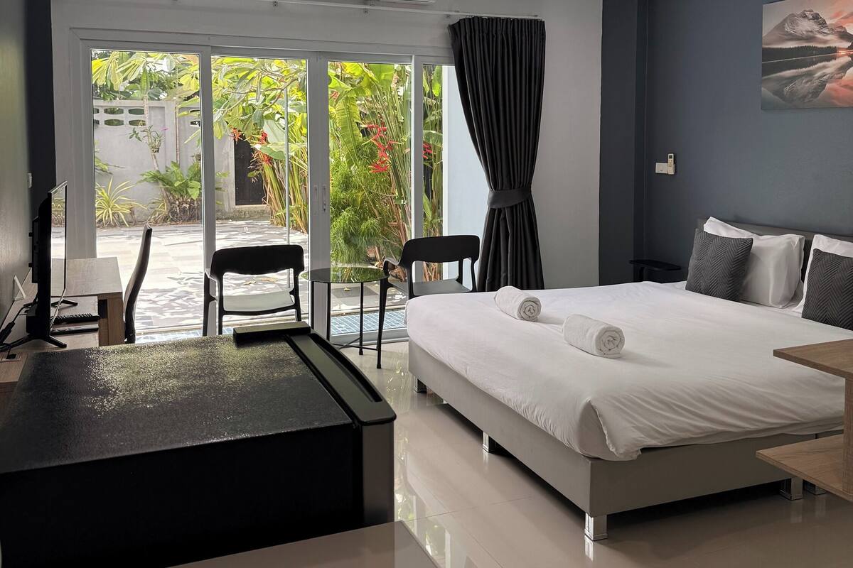 A modern hotel room with a bed, desk, chairs, and a view of greenery through large windows.
