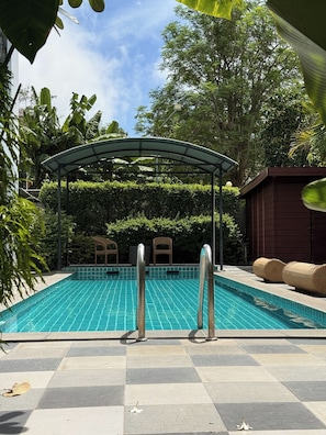 A swimming pool with a covered seating area, surrounded by greenery and a tiled deck.
