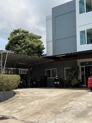 A building with a covered parking area, several motorcycles, and a large tree.