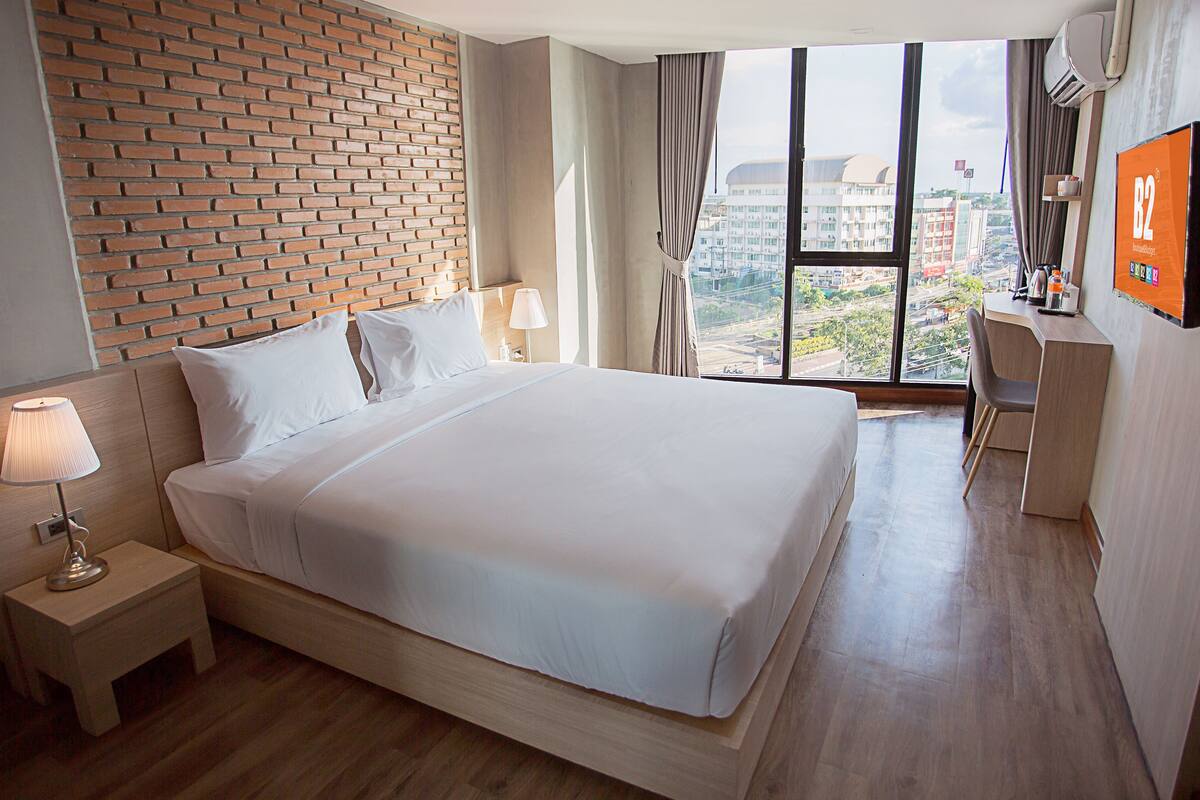 Une chambre d’hôtel avec un grand lit, un bureau, une télévision et une vue sur la ville grâce à de grandes fenêtres.