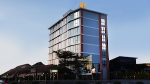 Un hotel de varios pisos con un llamativo letrero 'B2' en la parte superior, que presenta un diseño arquitectónico moderno con grandes ventanales y una combinación de paneles exteriores rojos y azules.