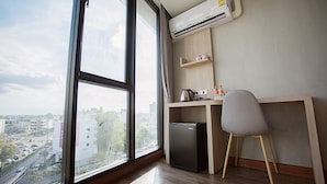 Una habitación con un ventanal grande, un escritorio con silla, un refrigerador pequeño y un estante con botellas y una tetera.