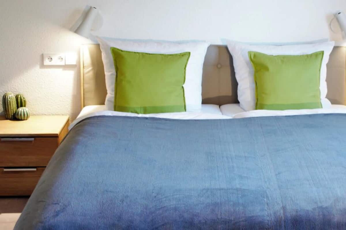 A bed with a blue blanket, two green pillows, and a wooden nightstand with a cactus.