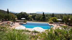 Zona de piscina con sillas de descanso y sombrillas, rodeada de vegetación y vistas a montañas lejanas.