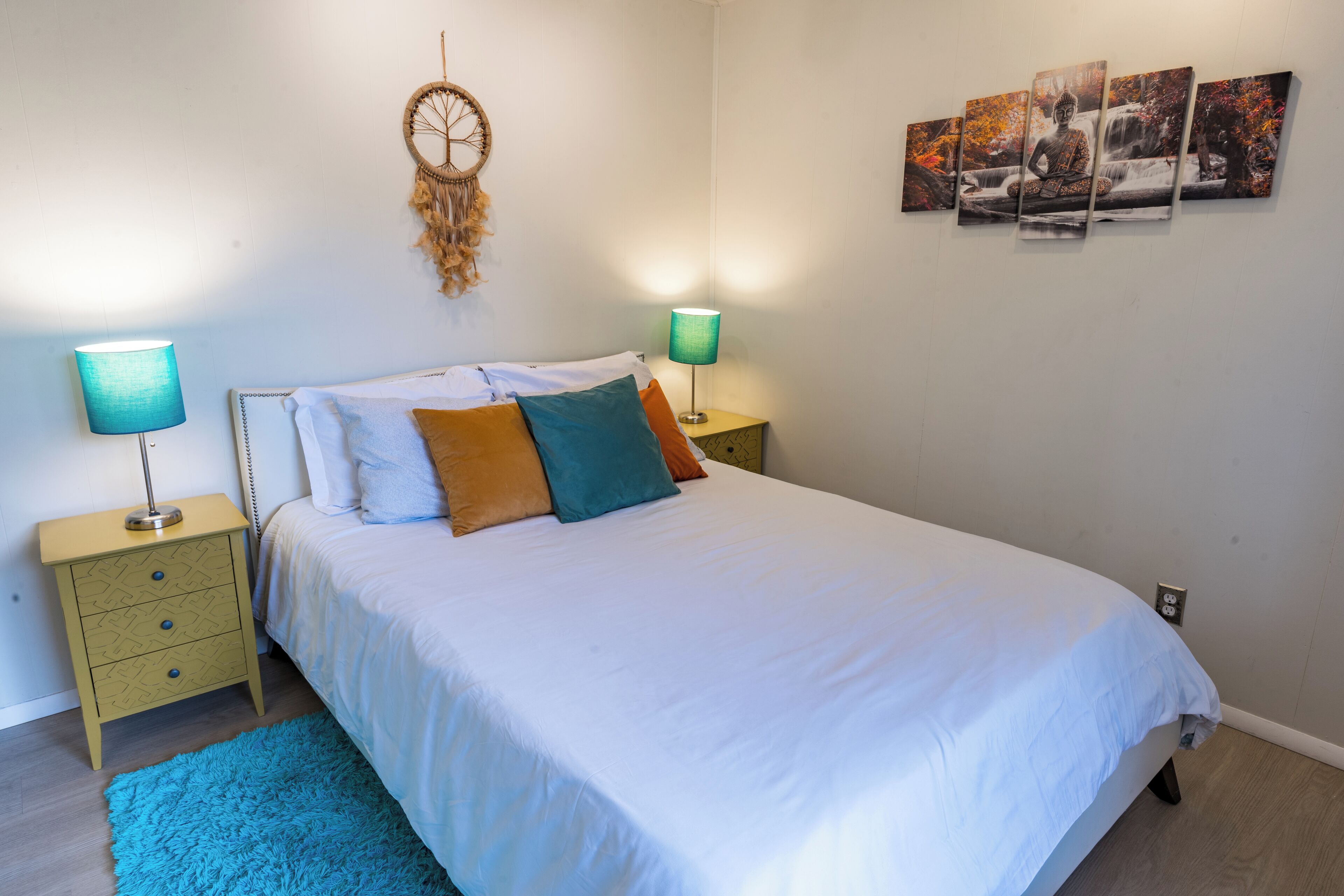 Una cama bien hecha con edredón blanco y varios almohadones, dos mesitas de noche con lámparas, un atrapasueños en la pared y una serie de cuadros enmarcados.
