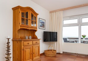 Ein Holzkabinent mit Glastüren, ein Fernseher an der Wand, ein Fenster mit Vorhängen und ein Korbwäsche am Boden.