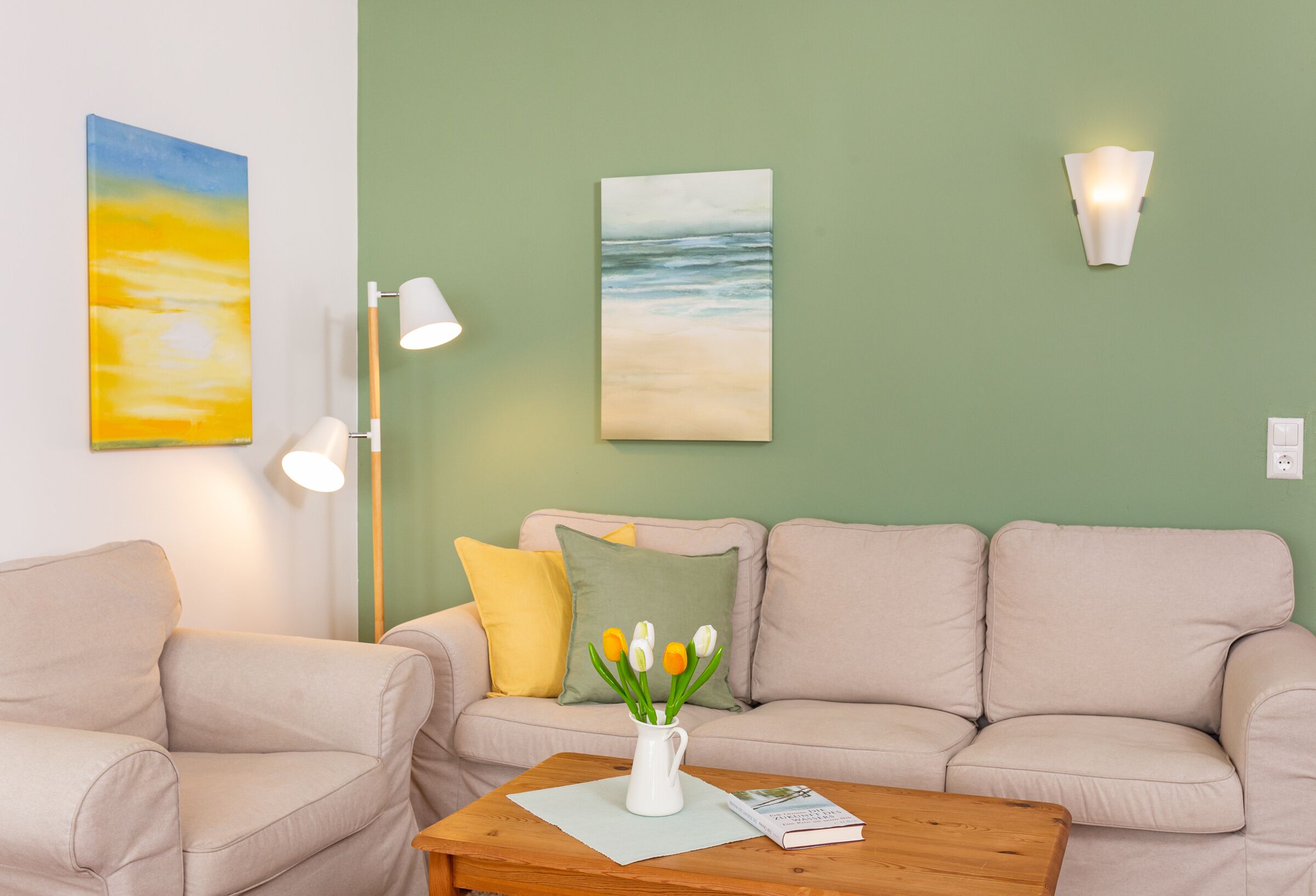 A living room with a beige sofa, a wooden coffee table with a vase of flowers, a lamp, and a painting on the wall.
