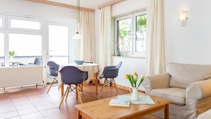 A modern living room with a beige sofa, a wooden coffee table, and a dining area with a white table and blue chairs. - Fantastic holiday apartment with beautiful views to the sea, it is dog friendly (Wittdün)