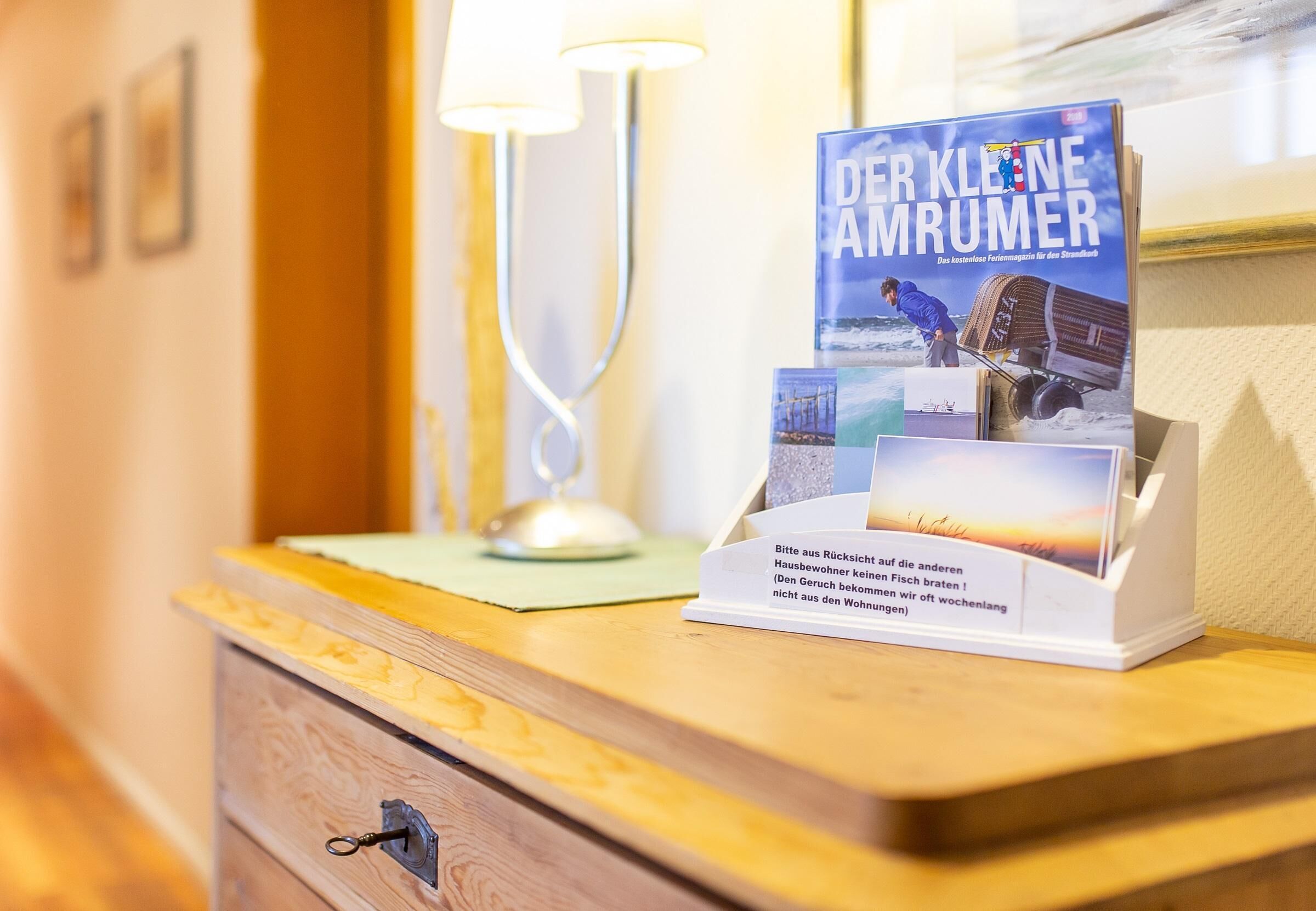 A hotel reception desk with a brochure stand displaying a magazine titled 'DER KLENE AMRUMER' and a small sign with a message in German.