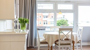 A dining area with a round table, white chairs, and a view of buildings through large windows. - Wonderful holiday flat with ocean view to the island Amrum - Seaview, Dogfriendl (Wittdün)