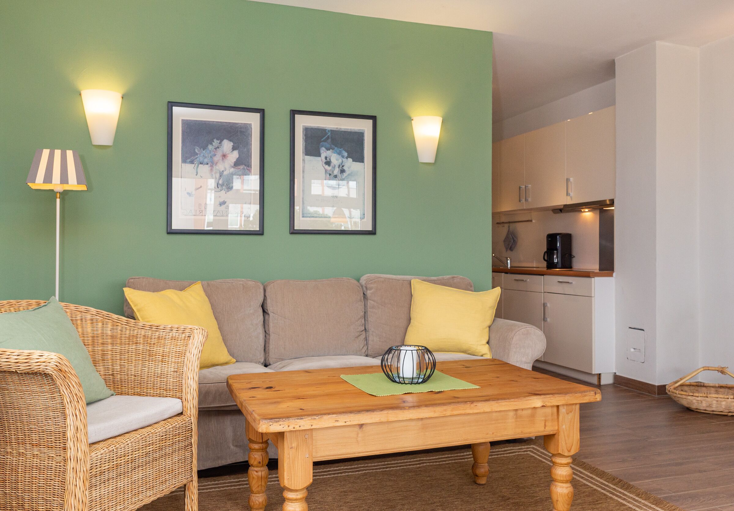 Sala de estar com sofá, mesa de centro de madeira e cadeira de vime. Há duas fotos emolduradas na parede e um abajur no chão.