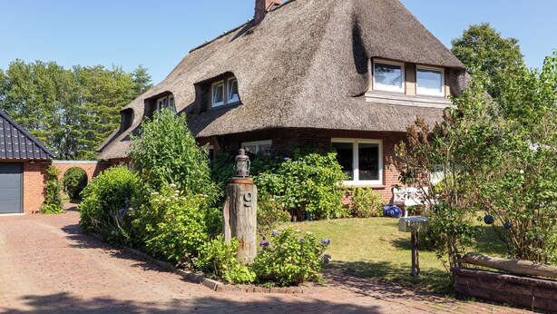 Une maison avec un toit de chaume, entourée d’un jardin bien entretenu où poussent différentes espèces de plantes, et une boîte aux lettres.