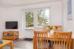 Une pièce avec une table à manger en bois, deux chaises, un téléviseur fixé au mur et une fenêtre donnant sur des arbres.