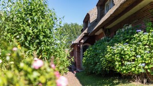 Une maison traditionnelle en briques, dotée d’un toit de chaume, entourée d’une végétation luxuriante et de plantes en fleurs.