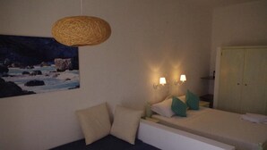 A bedroom with a bed, a sofa, a wall-mounted lamp, and a painting of a rocky landscape. - Hotel Esperos (Skiathos)