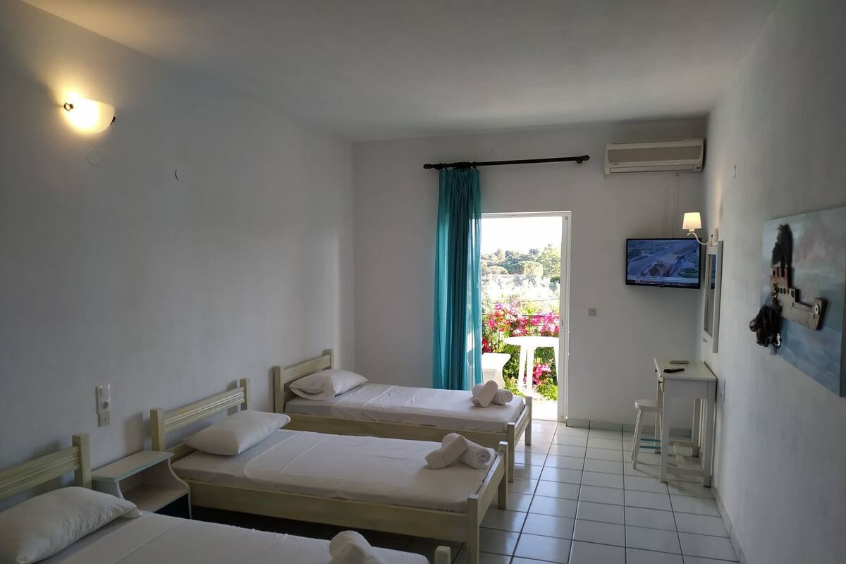 A hotel room with two single beds, a small table, a TV, and a balcony with a view of greenery and flowers.