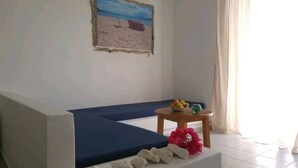 A room with a blue sofa, a small round table with fruits, and a painting of a beach scene. - Hotel Esperos (Skiathos)