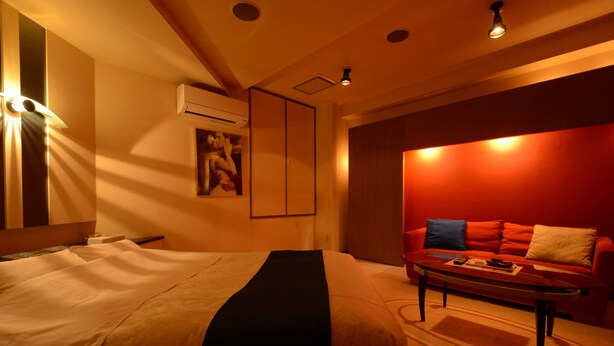 Habitación de hotel con cama, sofá rojo, mesa de centro y unidad de aire acondicionado en la pared.