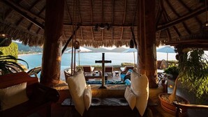 A thatched-roof veranda with a view of the sea and mountains, two white cushioned chairs, and a table with a cross in the center. - Steps from the beach, far from routine. An experience beyond the ordinary (Playa la ropa)