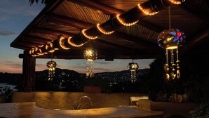 A covered outdoor area with a hot tub, decorative hanging lights, and a view of a distant cityscape at dusk. - Steps from the beach, far from routine. An experience beyond the ordinary (Playa la ropa)