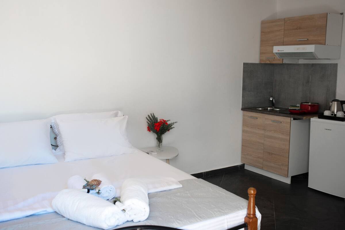 Hotelkamer met een bed, een klein nachtkastje met een bloemenvaas, een kitchenette met een fornuis en een koelkast, en een houten kastje boven de wastafel.